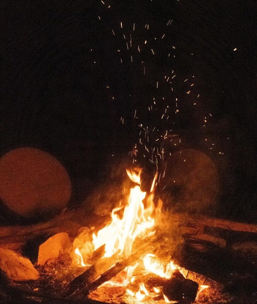les flammes d'un Feu Sacré crépitent dans la nuit. on voit le bois au centre du cercle de feu et les tambours chamaniques des stagiaires posés au sol autour de celui ci. cela dégage une ambiance sereine et mystique